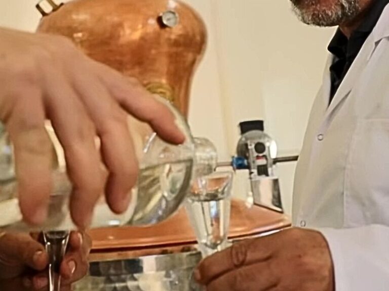 osoba sipa osobi rakiju u čašicu, u pozadini metalno bure za rakiju